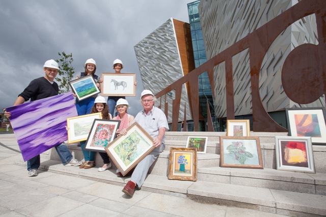 A group of people holding various sized artwork, the Titanic Building in Belfast can be seen in the background