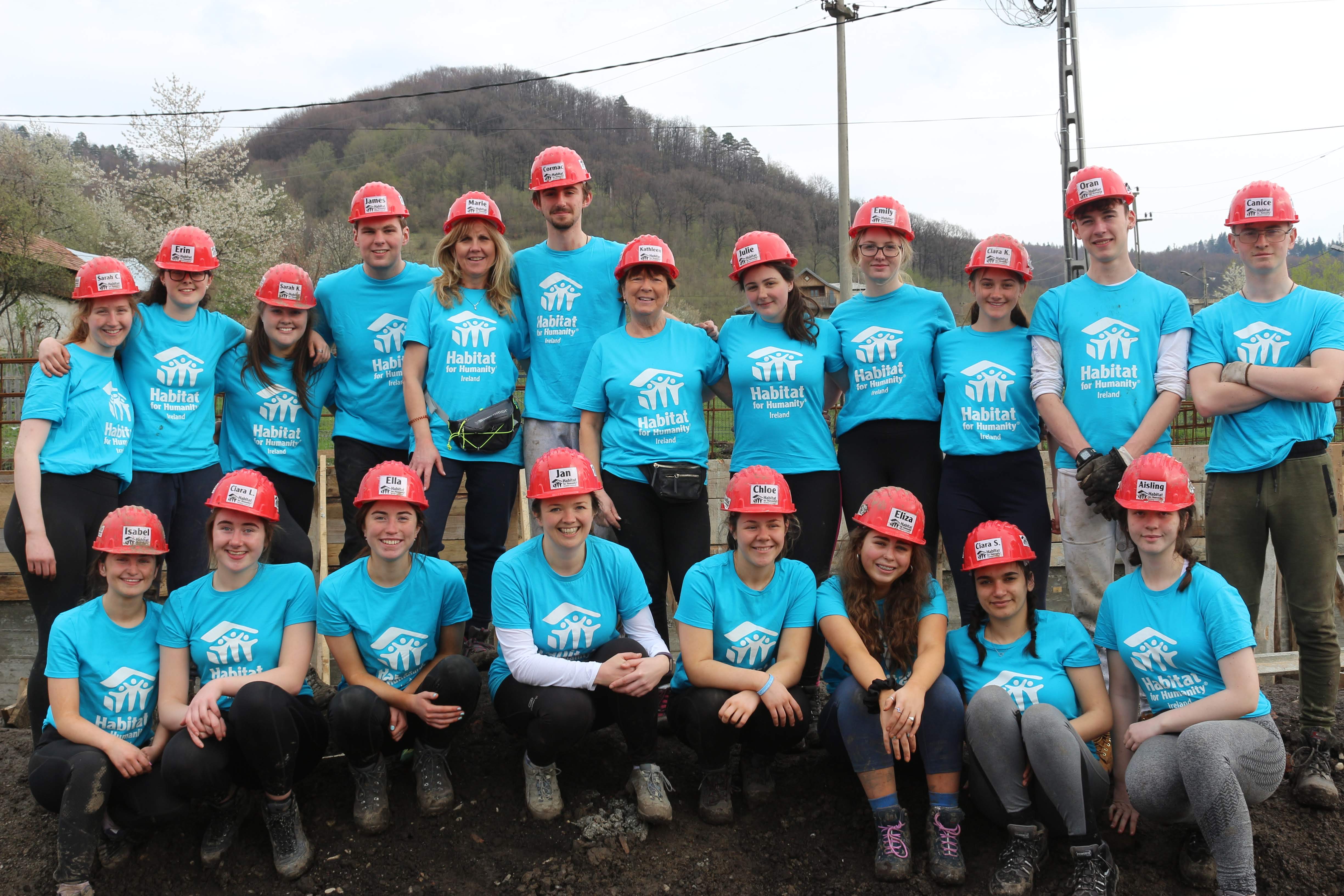 A group of volunteers, all in blue Habitat t-shirts and red hard hats, smile into the camera on sire in Romania