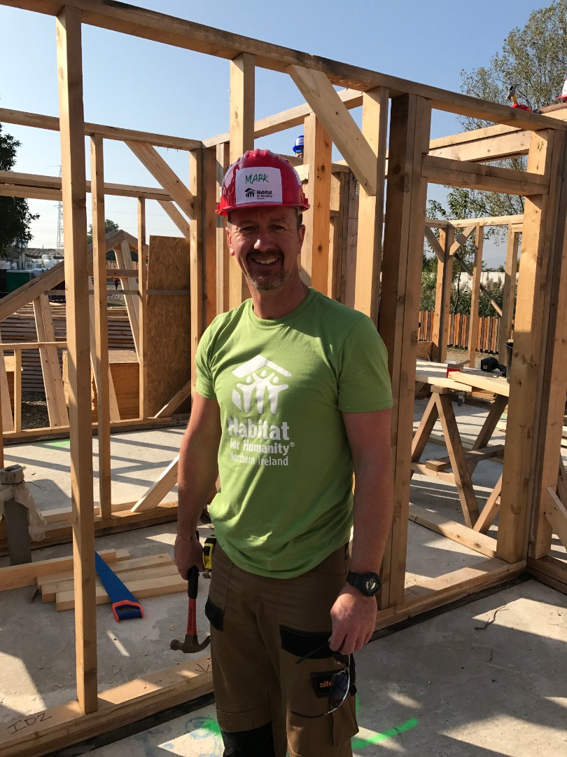 Mark Emerson wears a green Habitat t-shirt and red hard hat, with house frames in the background