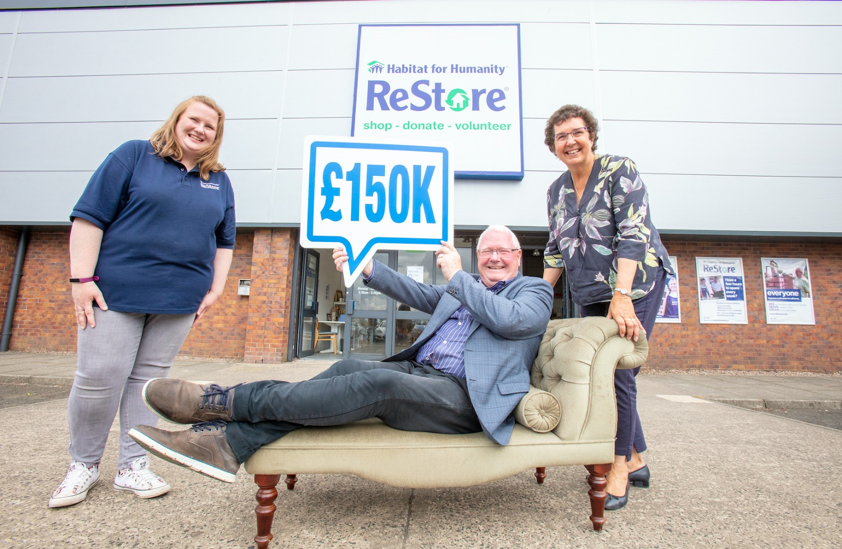 Staff from Habitat and the Gallaher Trust pose for a photo. One man is sitting on a chaise lounge, holding a sign showing a speech bubble with "£150k" written on it. The group are posing in front of the ReStore in Ballymena