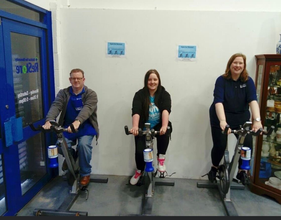 Three ReStore Ballymena staff/volunteers in-store on spin bikes, all smiling at the camera
