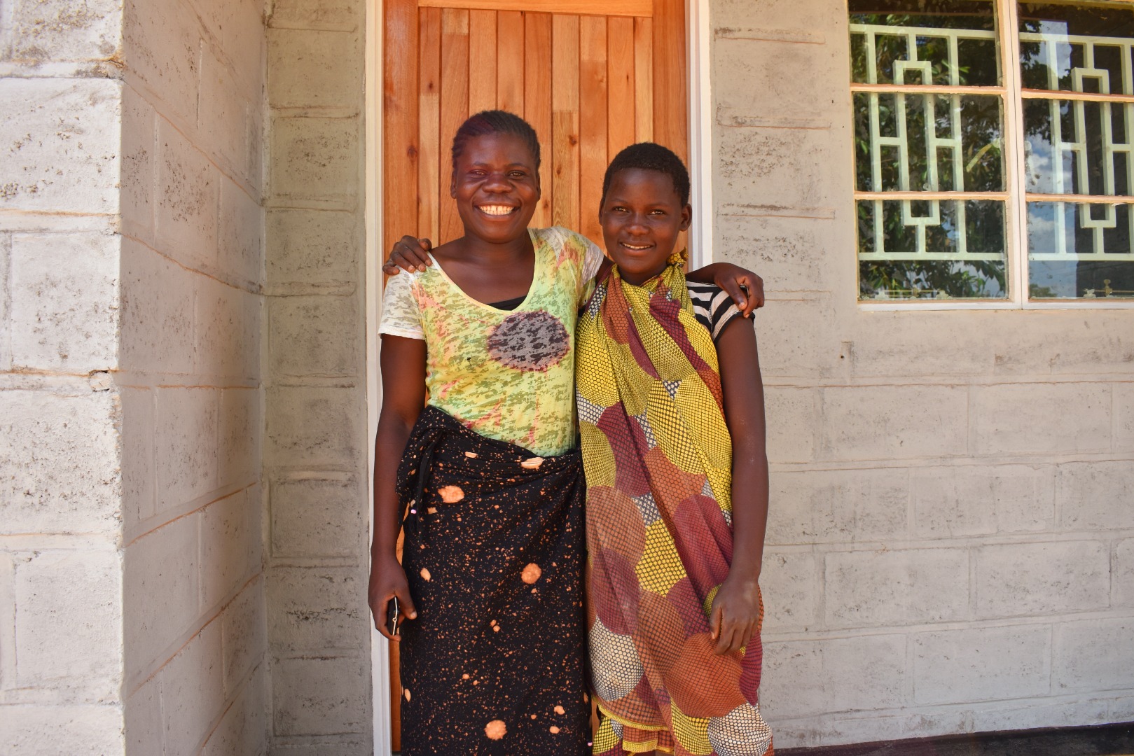 Chikondi and her sister stand, smiling, outside their Habitat home in Malawi. The sisters are both wearing colourful clothes and have their arms around each other.